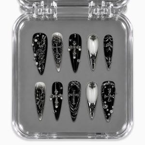 Peut inclure: Un ensemble de dix faux ongles en forme de stylet présentés dans un boîtier en plastique transparent. Les ongles arborent une palette de couleurs noir et blanc avec des emblèmes de croix métalliques argentés, des textures en relief et des perles.
