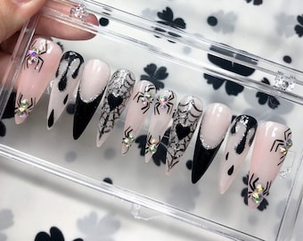 Könnte beinhalten: Ein Set aus zehn langen, spitz zulaufenden Kunstnägeln mit Halloween-Motiv. Zu den Designs gehören schwarze Spinnennetze, tropfende schwarze Farbe und glitzernde Spinnenakzente auf einem blassrosa Untergrund. Die Nägel werden in einer transparenten Kunststoffbox präsentiert.