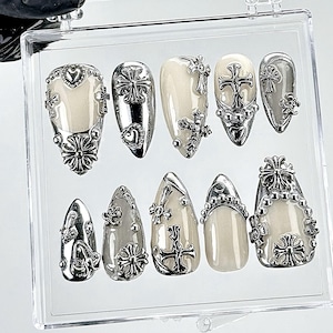 May include: A set of ten artificial nails in a clear plastic case. The nails are various shapes and feature a cream-coloured base with silver-coloured cross and floral embellishments. Some nails have heart-shaped accents.