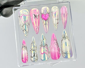 May include: A set of ten iridescent, long, pointed artificial nails. Each nail features a unique design, including hearts, butterflies, and silver embellishments. The nails are displayed in a clear plastic case.