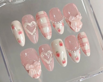 May include: A set of ten artificial nails in various designs. The nails feature a light pink color, with some adorned with white plaid patterns, strawberry designs, and heart-shaped accents. Some nails have a bow or a line of small, silver-colored beads.