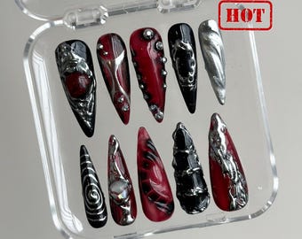Op de afbeelding: Een set van tien lange, puntige valse nagels in een doorzichtige plastic doos. De nagels hebben verschillende ontwerpen, waaronder zilveren versieringen, strass-steentjes en wervelende patronen. Kleuren zijn zwart, rood en zilver. Het woord "HOT" is rood gestempeld.