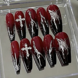 Puede incluir: Conjunto de diez u&ntilde;as postizas de color rojo y negro degradado con dise&ntilde;os de cruz y mariposa plateadas.