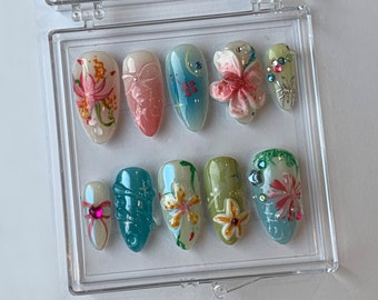 May include: A set of ten artificial nails in a clear plastic case. The nails feature various designs, including floral patterns, bows, and embellishments. Colors include pink, blue, green, and white. Some nails have 3D floral accents and rhinestones.
