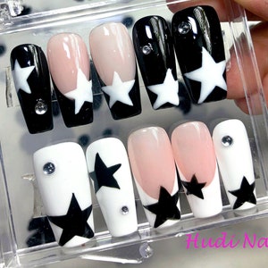 May include: A set of press-on nails in a clear plastic case. The nails feature a variety of designs, including black and white star patterns, a nude pink base with a white star, and a clear gem. The text "Hudi Nails" is visible.