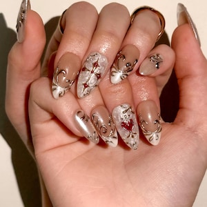 May include: Almond-shaped press-on nails featuring a nude base with intricate silver metallic filigree swirls, raised silver cross accents with small red gems, and a prominent red heart-shaped gem on the ring finger nail.