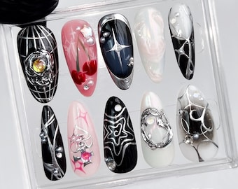 May include: A set of ten artificial nails with various designs. The nails feature a mix of black, pink, and white bases, adorned with silver embellishments, cherries, and starburst patterns. Some nails have a glossy finish, while others have a matte appearance.