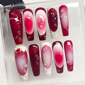 May include: A set of ten artificial nails in a clear plastic case. The nails are a deep red with a gradient effect, transitioning to a lighter pink and white. Each nail features a water droplet design.
