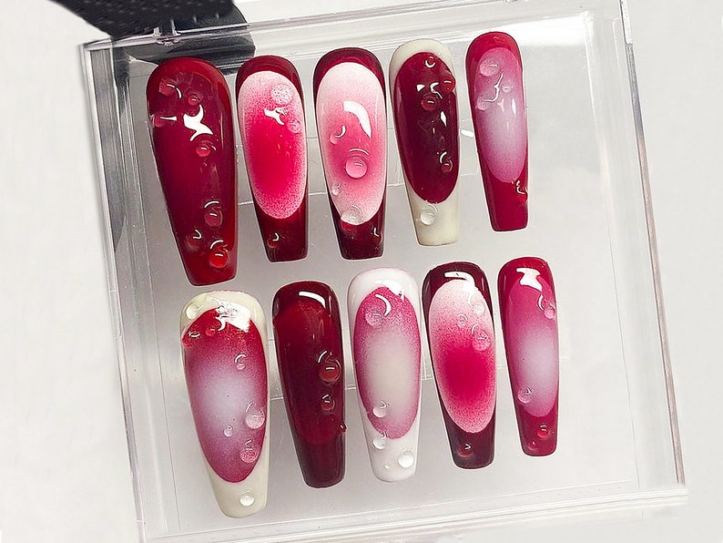 May include: A set of ten artificial nails in a clear plastic case. The nails are a deep red with a gradient effect, transitioning to a lighter pink and white. Each nail features a water droplet design.
