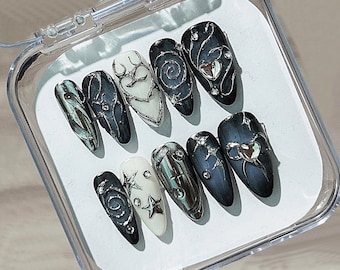 Op de afbeelding: Een set van tien kunstnagels in een doorzichtige plastic doos. De nagels hebben verschillende ontwerpen, waaronder zilveren wervelingen, sterren en hartvormige versieringen. De basiskleuren zijn zwart, wit en blauwgroen.
