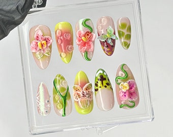 May include: A set of ten artificial nails with a variety of designs. The nails feature 3D floral and abstract patterns in shades of pink, yellow, green, and white. Some nails have a clear base with decorative elements.