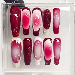 May include: A set of ten artificial nails in a clear plastic case. The nails are a deep red with a gradient effect, transitioning to a lighter pink in the center. Each nail features a droplet design. The tips are either white or red.
