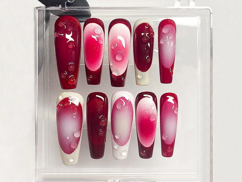 May include: A set of ten artificial nails in a clear plastic case. The nails are a deep red with a gradient effect, transitioning to a lighter pink in the center. Each nail features a droplet design. The tips are either white or red.