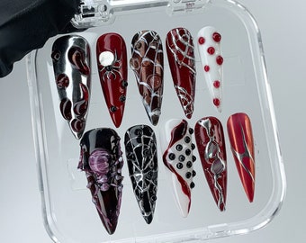 May include: A set of ten long, pointed false nails with various Halloween-themed designs. The nails feature colors like black, red, silver, and white, with spiderwebs, spiders, and abstract patterns. The nails are displayed in a clear plastic case.