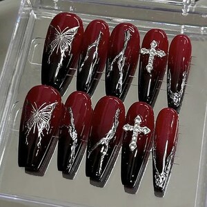 Puede incluir: Conjunto de diez u&ntilde;as postizas de color rojo y negro con dise&ntilde;os de mariposas y cruces plateados.