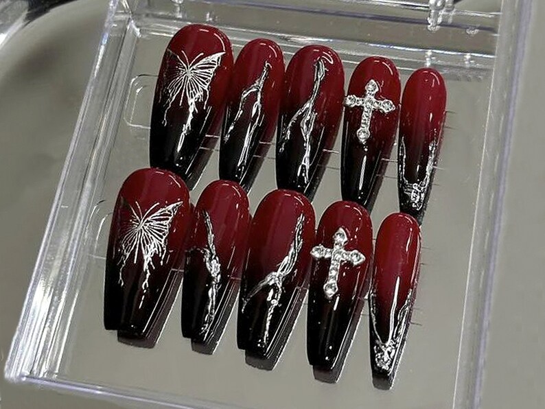 Puede incluir: Conjunto de diez u&ntilde;as postizas de color rojo y negro con dise&ntilde;os de mariposas y cruces plateados.