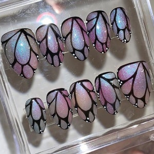 May include: A set of press-on nails with an iridescent butterfly wing design. The nails have a colour gradient of silver, purple, and blue, with black outlines and small white dots. They are presented in a clear plastic case.