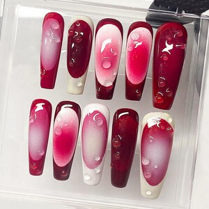 May include: A set of ten artificial nails in a clear plastic case. The nails are a deep red with a white and pink gradient design. Each nail has a water droplet design.