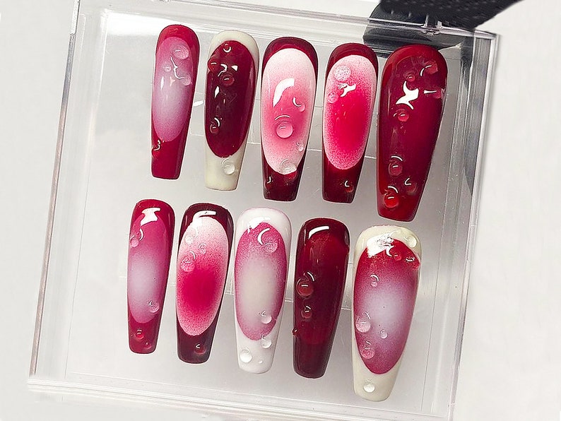 May include: A set of ten artificial nails in a clear plastic case. The nails are a deep red with a white and pink gradient design. Each nail has a water droplet design.