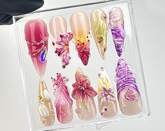 Könnte beinhalten: Ein Set aus zehn künstlichen Nägeln in einer transparenten Präsentationsbox. Die Nägel weisen eine Vielzahl von Designs auf, darunter florale Verzierungen, Strasssteine und Farbverläufe in Rosa, Pfirsich, Gelb und Lila. Die Nägel sind lang und spitz.