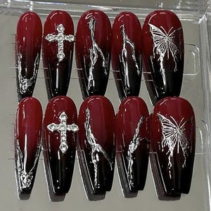 Puede incluir: Conjunto de diez u&ntilde;as postizas largas, de forma de ata&uacute;d, de color rojo y negro degradado con detalles plateados. Algunas u&ntilde;as tienen una cruz plateada, otras tienen una mariposa plateada.