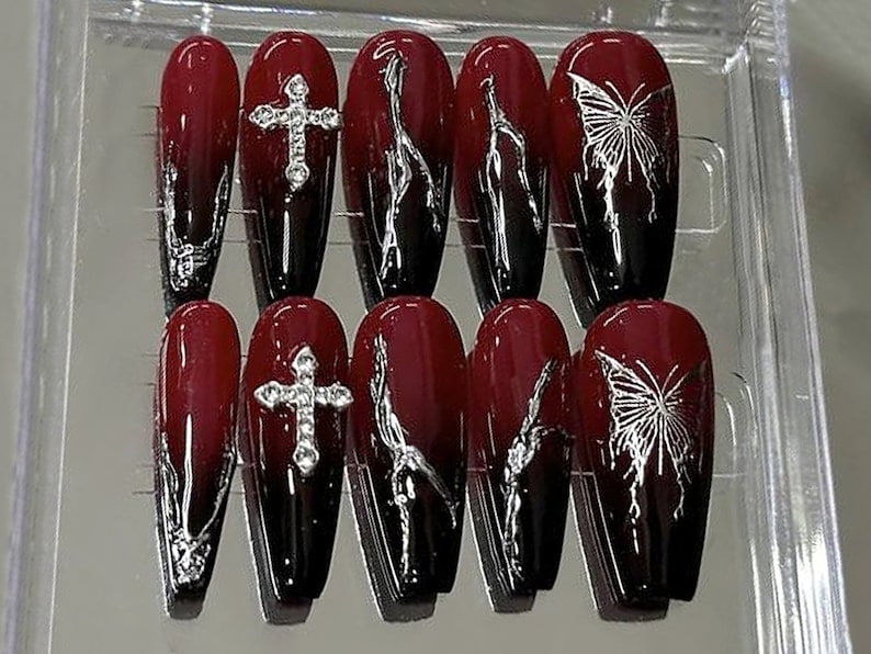 Puede incluir: Conjunto de diez u&ntilde;as postizas largas, de forma de ata&uacute;d, de color rojo y negro degradado con detalles plateados. Algunas u&ntilde;as tienen una cruz plateada, otras tienen una mariposa plateada.