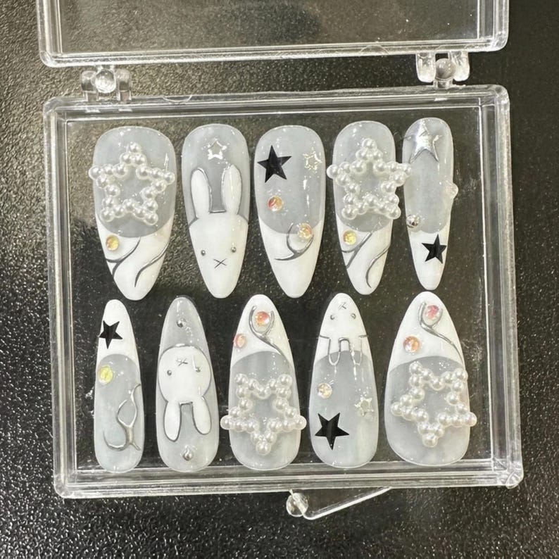 May include: A set of press-on nails in a clear plastic case. The nails feature a gray and white design with star and bunny accents. Some nails have pearl star embellishments and black star details.