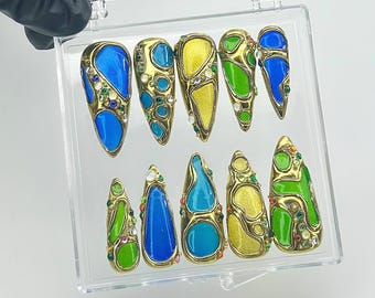 Op de afbeelding: Een set van tien sierlijke, goudkleurige nagelkunststukken. Elke nagel heeft een uniek ontwerp met levendig blauw, groen en geel emaille, geaccentueerd met kleine, kleurrijke edelstenen. Gepresenteerd in een doorzichtige plastic doos.