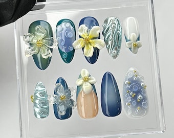 May include: A set of ten artificial nails in a clear plastic case. The nails feature various designs, including floral embellishments, swirls, and gradients in shades of blue, green, and white. Some nails have pearl accents.