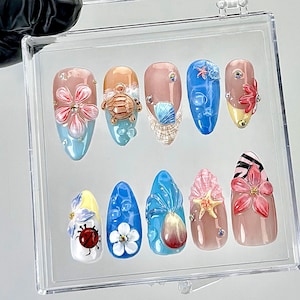 May include: A set of ten artificial nails in a clear plastic case. Each nail features a different design, including flowers, a turtle, seashells, and a starfish. The nails are painted in various colors, including pink, blue, and white.