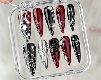 Op de afbeelding: Een set van tien lange, puntige valse nagels in een doorzichtige plastic doos. De nagels hebben verschillende ontwerpen, waaronder zilveren manen, sterren en abstracte patronen, met een kleurenpalet van zwart, bordeauxrood en zilver.