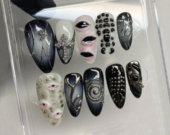Op de afbeelding: Een set van tien nepnagels met verschillende ontwerpen. De nagels hebben een mix van zwarte, witte en grijze kleuren met zilveren accenten. Sommige nagels hebben kruis-, oog- en abstracte ontwerpen.