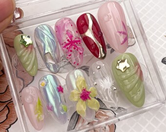 May include: A set of ten artificial nails in a clear plastic case. The nails feature various designs, including iridescent, floral, and abstract patterns. Colors include pink, green, red, and gold, with embellishments like rhinestones and star-shaped charms.