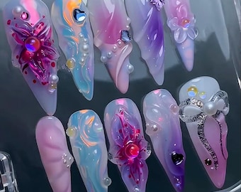 May include: A set of ten long, sculpted acrylic nails. Each nail features a unique design with iridescent colors, pearls, and floral or gem embellishments. The color palette includes pinks, purples, blues, and iridescent accents, creating a glamorous aesthetic.