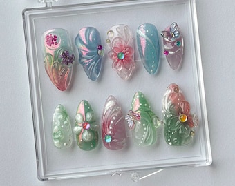 Könnte beinhalten: Ein Set aus zehn künstlichen Nägeln in einer transparenten Kunststoffbox. Die Nägel weisen eine Vielzahl von Designs auf, darunter florale und Schmetterlingsakzente, schillernde Farben und dekorative Edelsteine. Die Nägel sind oval geformt und variieren in der Farbe von Rosa bis Grün.