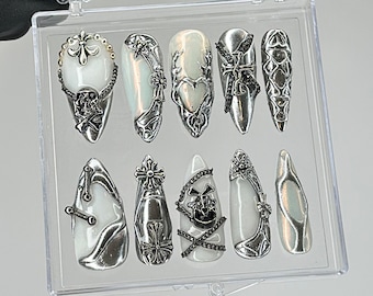 Op de afbeelding: Een set van tien kunstnagels met een witte basis en sierlijke zilverkleurige versieringen. Ontwerpen omvatten kruisen, schedels, kettingen en bloempatronen. De nagels worden gepresenteerd in een doorzichtige plastic doos.