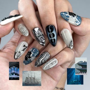 May include: A hand showcases a set of long, almond-shaped nail designs. The nails feature a variety of patterns, including silver swirls, geometric shapes, and floral accents in black and blue. Some nails have a glossy finish, while others are matte.