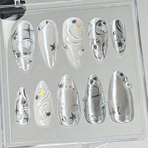 May include: A set of silver press-on nails with various designs. Some nails feature black swirls, stars, and clear rhinestones. Others have a pearlescent finish with yellow star accents. The nails are displayed in a clear plastic case.