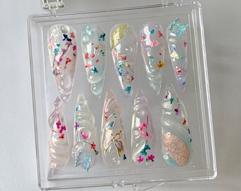 May include: A set of ten clear acrylic nails with various floral and butterfly designs. Each nail features embedded dried flowers, glitter, and 3D butterfly accents in shades of pink, blue, and gold, encased in a clear, glossy finish.
