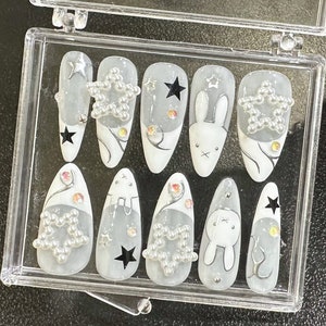 May include: A set of press-on nails in a clear plastic case. The nails feature a white and gray design with star and bunny accents. Some nails have star-shaped embellishments, and others have black star decals.