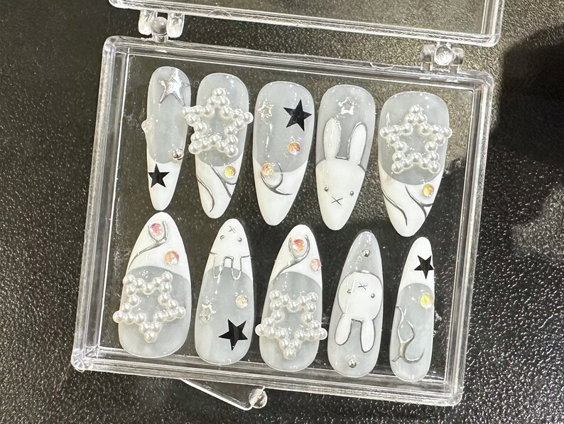 May include: A set of press-on nails in a clear plastic case. The nails feature a white and gray design with star and bunny accents. Some nails have star-shaped embellishments, and others have black star decals.