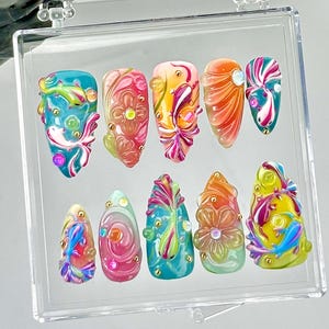 May include: A set of ten artificial nails in a clear display case. Each nail features a unique design with vibrant colours like teal, pink, orange, and yellow. The designs include floral and abstract patterns with gold accents.