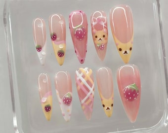 May include: A collection of ten artificial nails with diverse designs. The nails showcase a pale pink base with strawberry, bear, and plaid patterns. Some nails have a clear, glossy finish with small water droplet details.