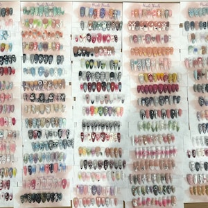 May include: A selection of false nails featuring diverse designs, such as floral patterns, cartoon characters, and glitter. The nails are presented on cards with the text "Hudi Nails".