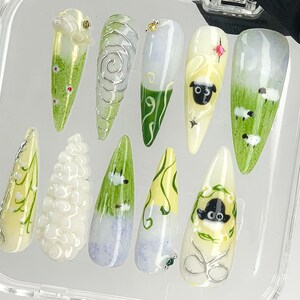Puede incluir: Un conjunto de diez u&ntilde;as artificiales con varios dise&ntilde;os. Las u&ntilde;as presentan una gama de colores, incluyendo blanco, verde y amarillo, con ilustraciones detalladas de ovejas, flores y patrones abstractos. Algunas u&ntilde;as tienen alambre de plata y detalles de gemas.