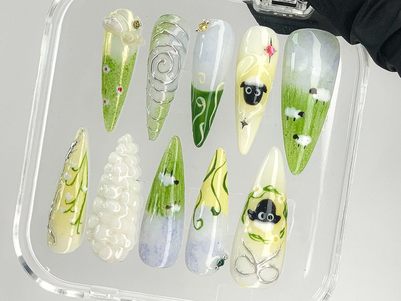 Puede incluir: Un conjunto de diez u&ntilde;as artificiales con varios dise&ntilde;os. Las u&ntilde;as presentan una gama de colores, incluyendo blanco, verde y amarillo, con ilustraciones detalladas de ovejas, flores y patrones abstractos. Algunas u&ntilde;as tienen alambre de plata y detalles de gemas.