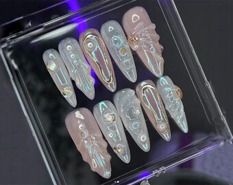 Könnte beinhalten: Ein Set aus langen, mandelförmigen Kunstnägeln. Die Nägel sind hellrosa mit schillernden Akzenten und goldfarbenen Verzierungen. Einige Nägel weisen ein strukturiertes Design auf, während andere eine glatte Oberfläche haben. Die Nägel sind in einem transparenten Kunststoffbehälter angeordnet.