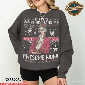 Peut inclure: Sweat-shirt gris anthracite avec l'inscription "OH IT'S CHRISTMAS AWESOME WOW !" et un motif d'une personne en manteau et chapeau rouge. Le design comprend des étoiles et des flocons de neige.