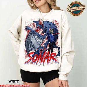 Puede incluir: Sudadera blanca con un gráfico de una criatura parecida a un murciélago y la palabra "SONAR" en rojo y azul. El diseño incluye un murciélago con grandes alas y una figura con traje. Cuello redondo, mangas largas.