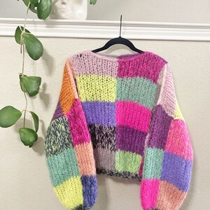 Puede incluir: Un suéter de punto de patchwork colorido con un corte holgado y mangas largas. El suéter está hecho de un hilo suave y peludo y presenta una variedad de colores, incluyendo rosa, morado, verde, amarillo y naranja.
